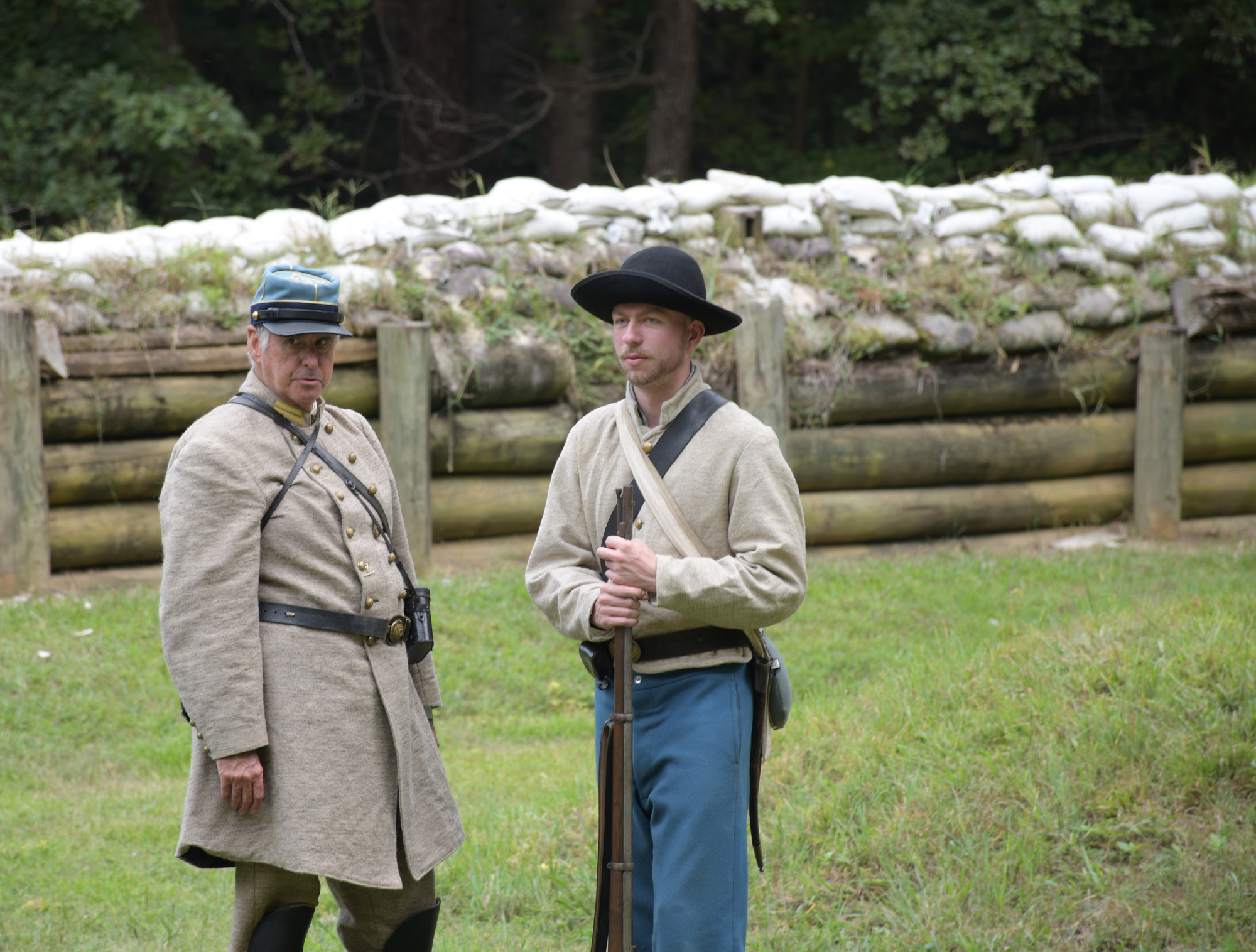 Two white men wearing Confederate uniforms one an officer and the other an enlisted man holding a rifle stand next to each other. A wooden retaining wall supports earthworks topped with sandbags in the background.