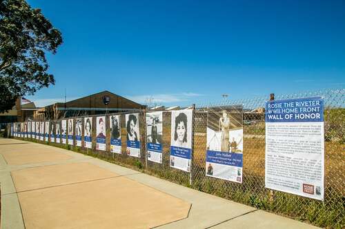 Multiple banners with historic and modern photos of home front workers, line a fence along a sidewalk. The park Visitor Education Center, a brick building with an NPS shield sign can be see in the background. 