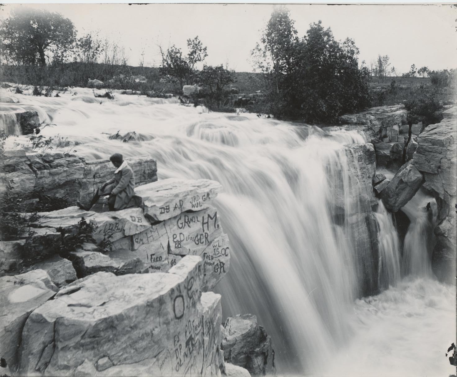 Black and white photo of a waterfall, graffiti on the rocks, and a woman sitting next to the edge