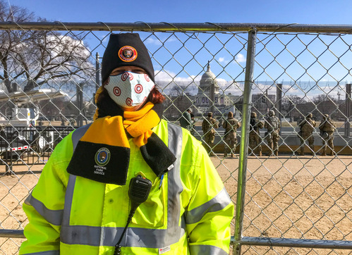A National Park Service volunteer wearing a face mask and radio standing next to a fence with the U.S. Capitol building in the distance