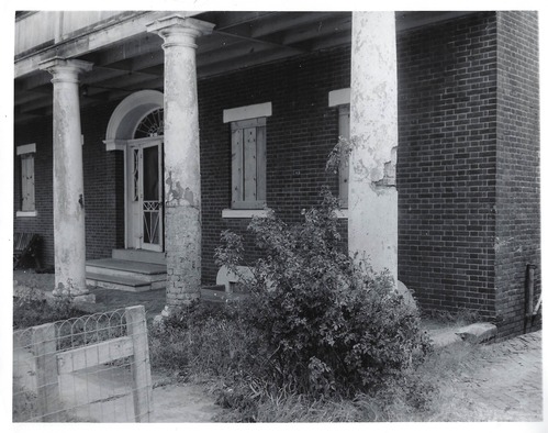 The south side of the Officer's Quarters.  Columns are in various states from peeled paint to the brick work under the "cement" overlay.  The windows are shuttered in the closed position, and a small shrub with a wood and wire fence is in the forefront of the picture.