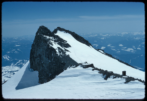 A string of buildings along a narrow rocky ridge that juts up out of a snowy slope high above the surrounding valleys.