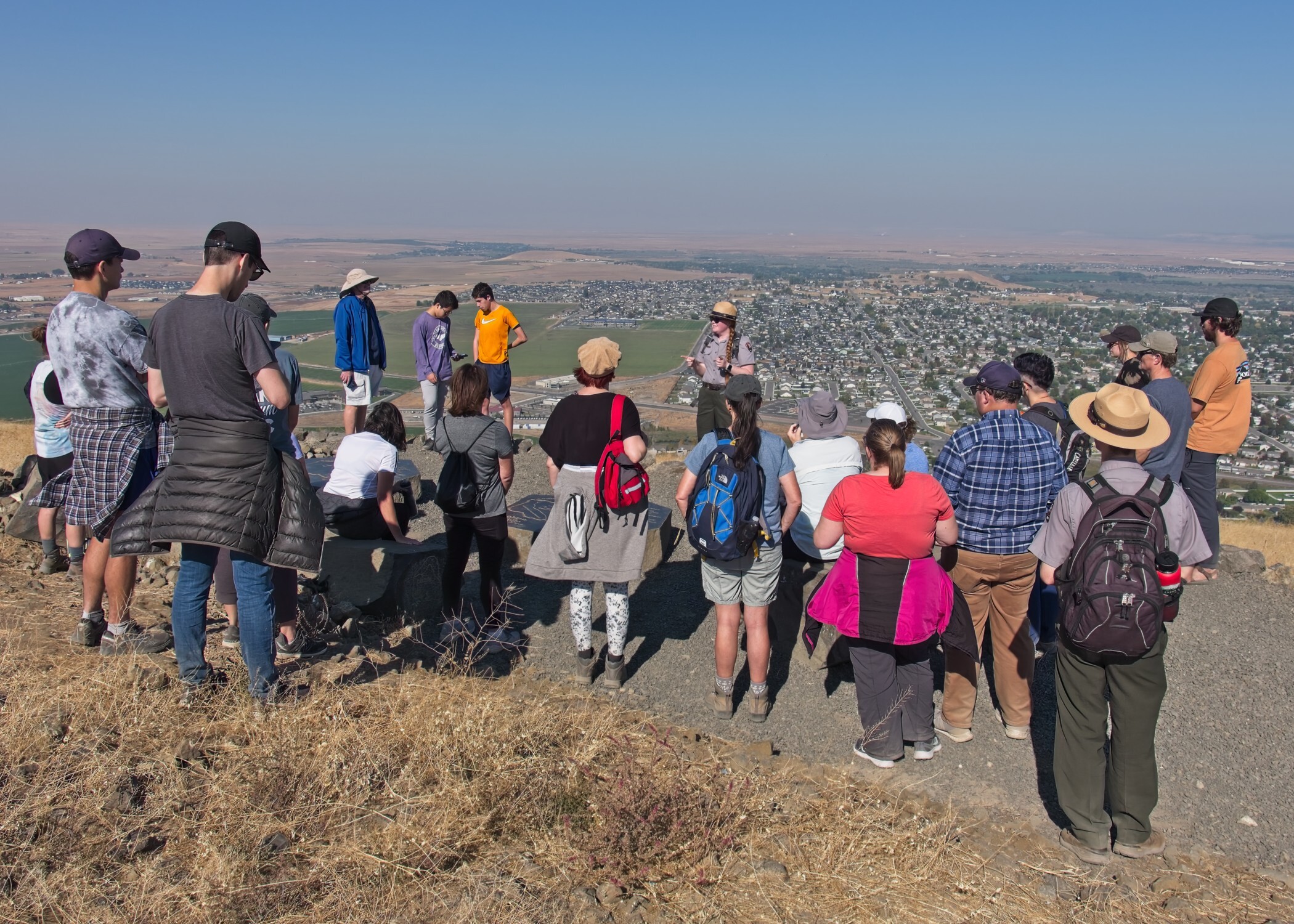 Three uniformed rangers stand surrounded by about fifty participants in a gravel lot trailhead with dried grass fields extending to the horizon.