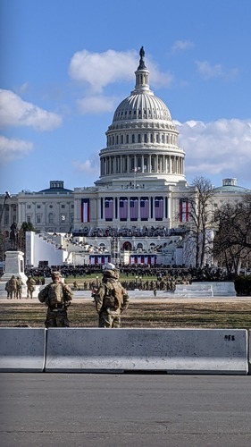 U.S. military standing watching the U.S. Capitol building, which has a large crowd gathered on its various levels and is decorated with large U.S. flags and red, white, and blue banners.