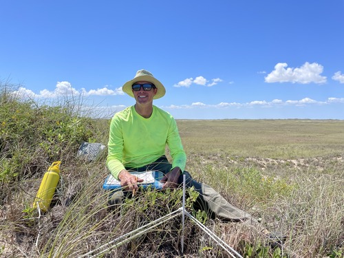 A smiling field crew member sitting in a grassland and holding a clipboard for recording names of plants 