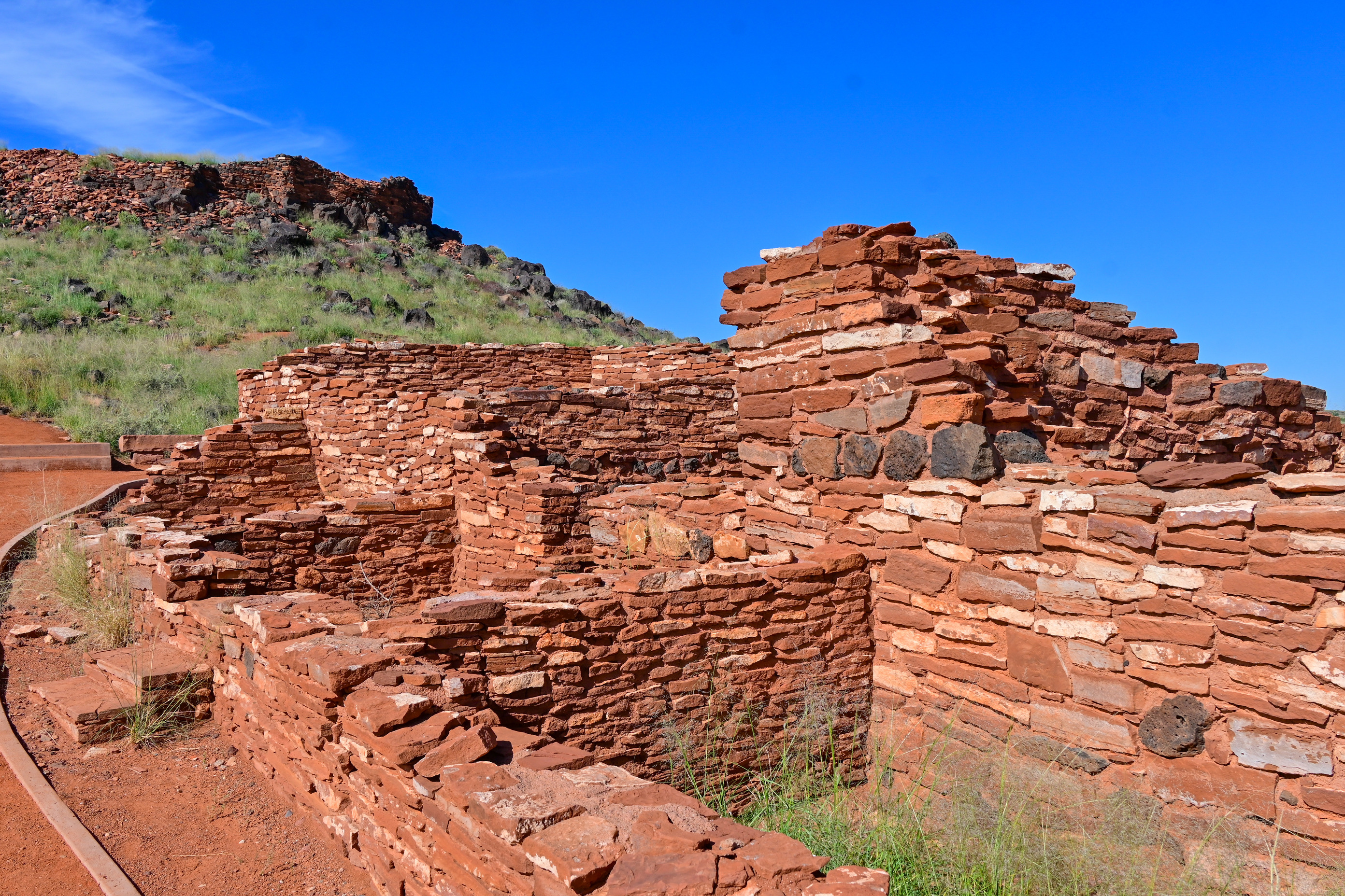 A small pueblo made of red sandstone sits next to a red sand path.  A second pueblo is visible above it on a hill.  