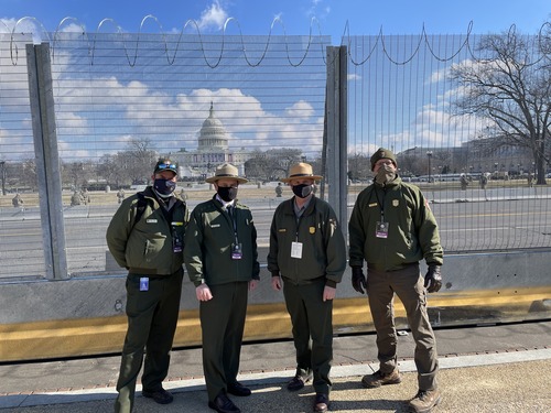Four uniformed National Park Service employees next to a fence with the U.S. Capitol building in the distance