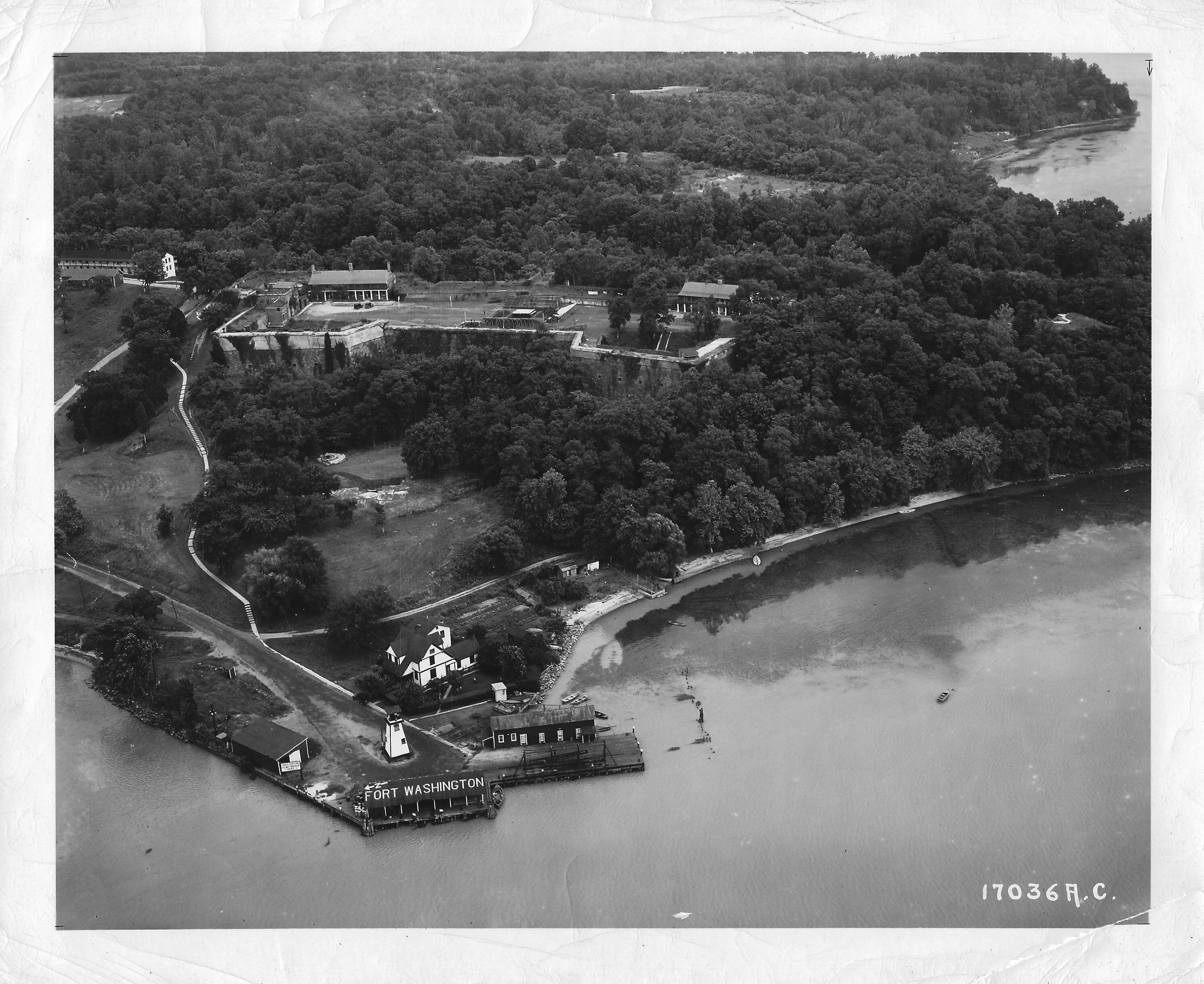 Photo shows the dock and lighthouse area.  The lighthouse keepers quarters is behind the lighthouse.  The fort is surrounded by heavy tree coverage, with several trees growing out of the southwest demi-bastion.  A small area of PIscataway Creek can be seen to the right of the photo.