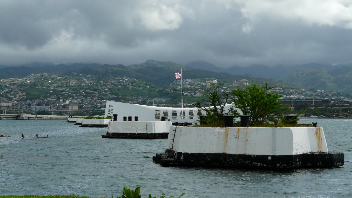 The elongated design and American flag of the USS Arizona Memorial is visible beyond a mooring quay, a structure built in the water for securing battleships. Cloud-shadowed hills rise in the distance on the other side of the water. 