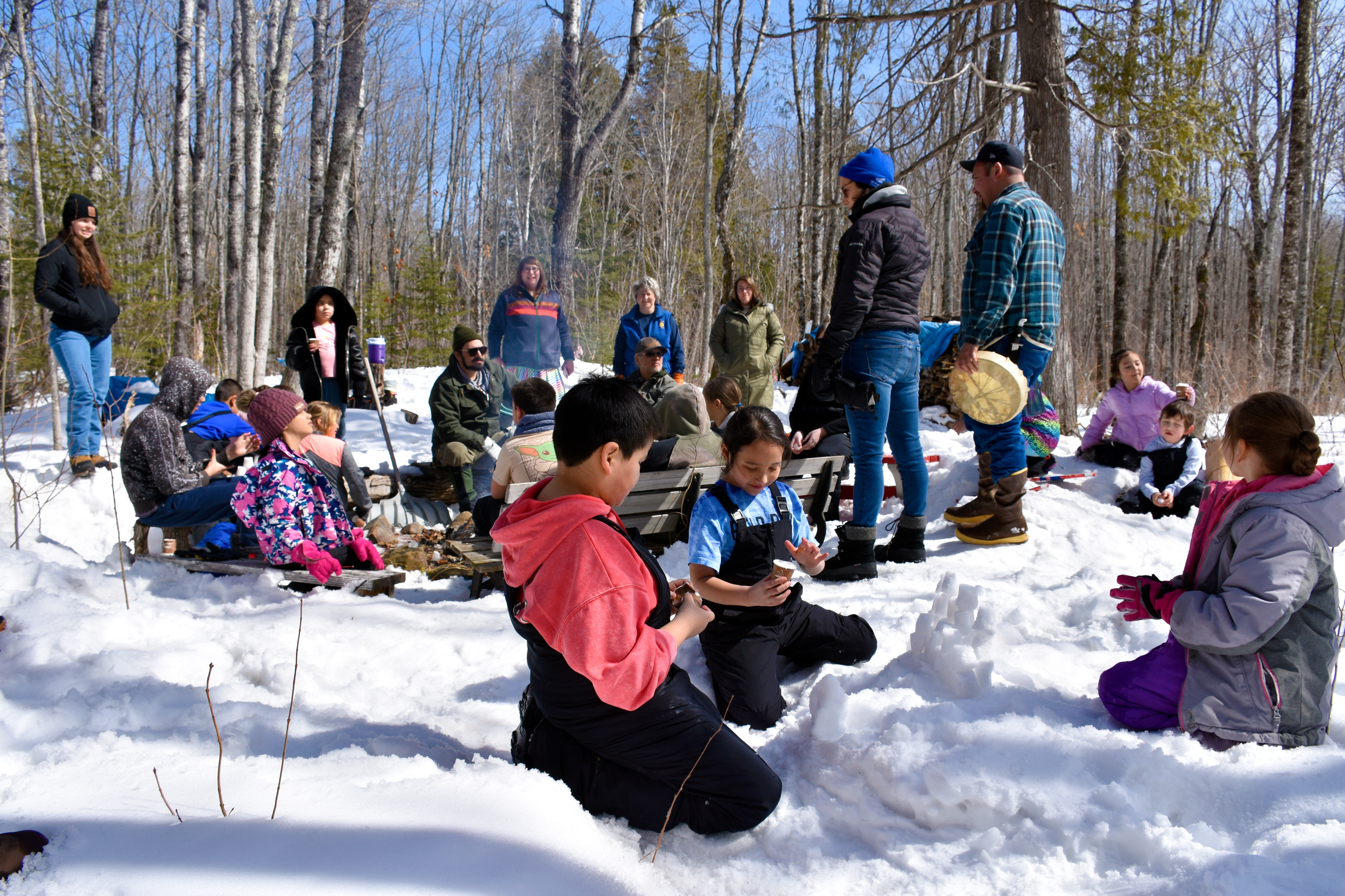 Children make a snow castle in the foreground in front of a group of children and adults standing and sitting on benches.