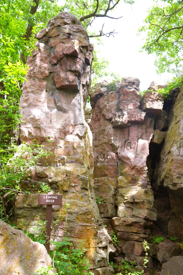 Two vertical rock pillars, with the left one a little closer and with a brown sign with white block letters in front labeling it as, "LEAPING ROCK" 
There are branches and leaves framing the left and right, with bright sky showing above the rock column to the right. 