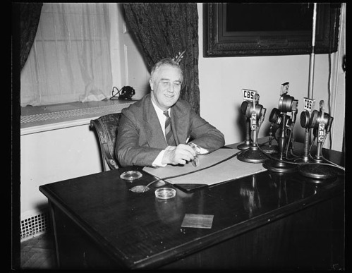 A man is seated behind an official looking desk in a suit. He is smiling widely with four antiquated microphones sitting on the desk and pointed at him at the right of the image. A desk mat and ashtray are the only other objects on the desk. Behind the man is a window with a deep sill, heavy drapery and the corner of an ornate picture frame. He is holding a pair of glasses in his right hand on top of the desk.