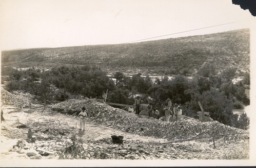 Black and white photo of a man standing in front of and several men digging rock on top of a large berm of rock parallel to a river