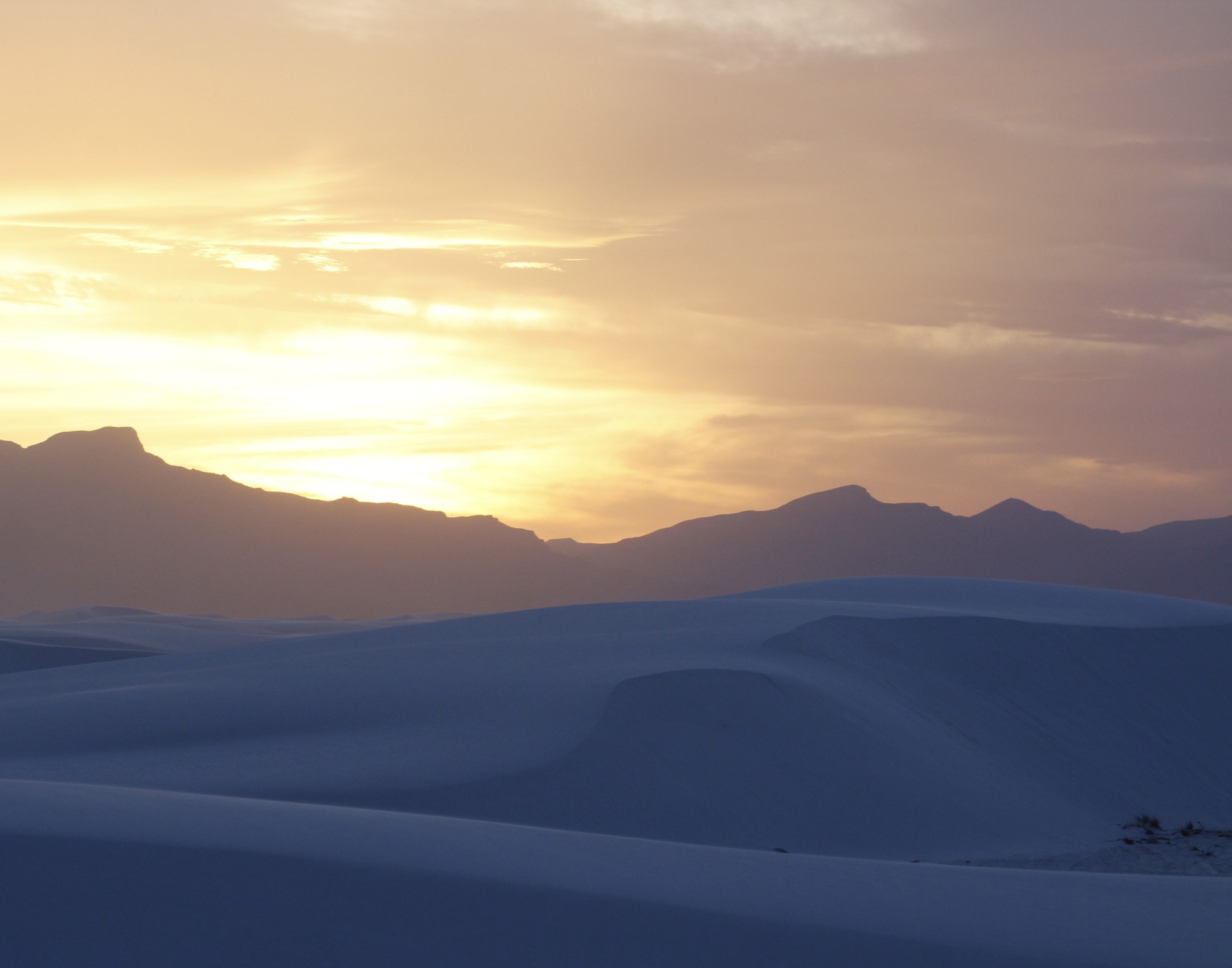 Tall rolling hills of sand dunes at the base of rugged mountains spanning the horizon of the image under a barely lit sunset sky. 