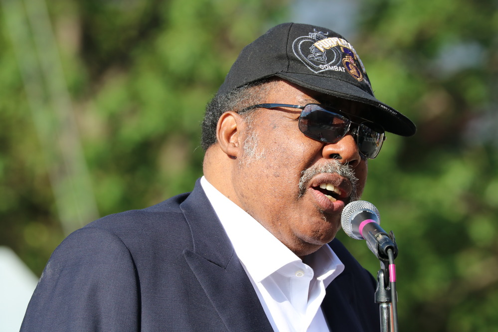 man seen from chest up wearing hat and sunglasses speaking into microphone