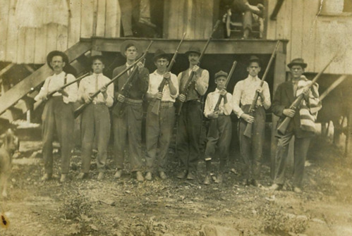 Eight men are posed in a line, standing in front of a raised, frame porch. They are coal miners and each man holds a rifle with the barrel point up to the right of the image. One of the miners looks eerily like a young teenage boy.