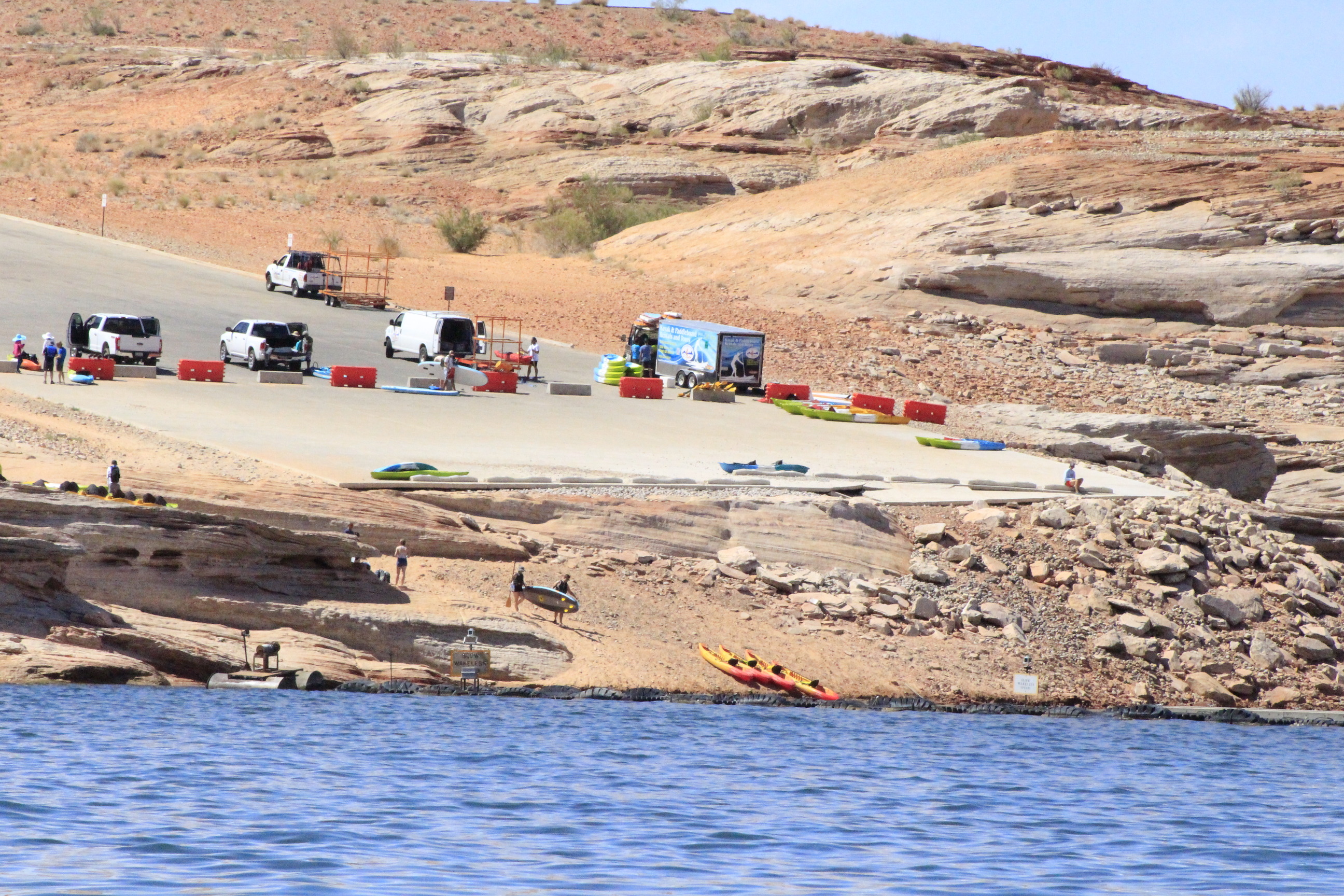 Trucks unloading kayaks on a barricaded launch ramp as visitors descend a rocky shoreline.