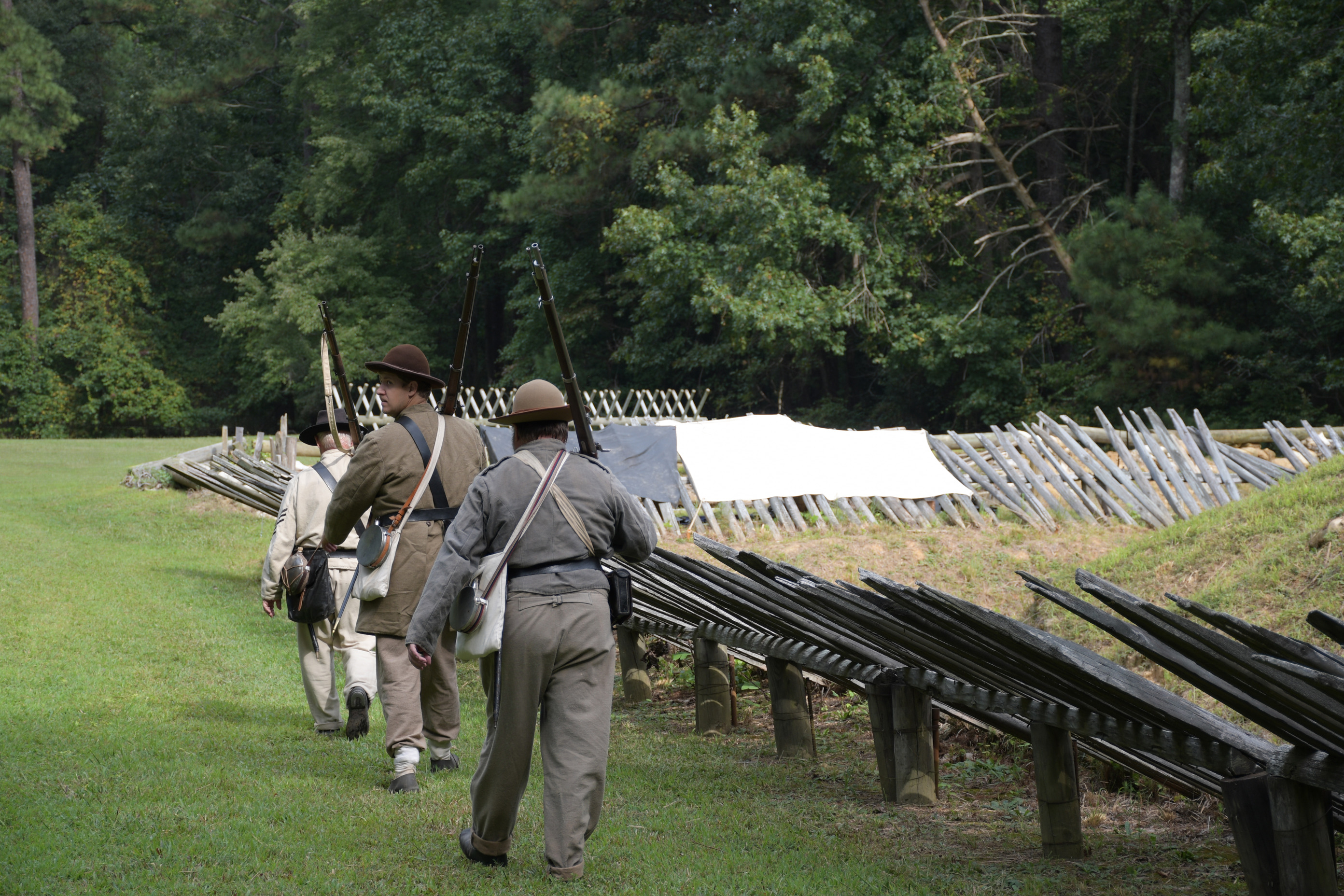 The three white men in Confederate uniforms carrying rifles walk past wooden palisades.