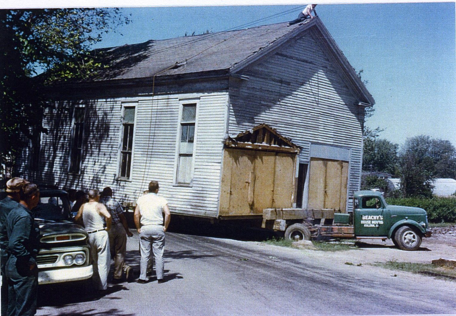 A green pickup truck tows a trailer carrying a long wood frame building with tall windows and a gabled roof.
