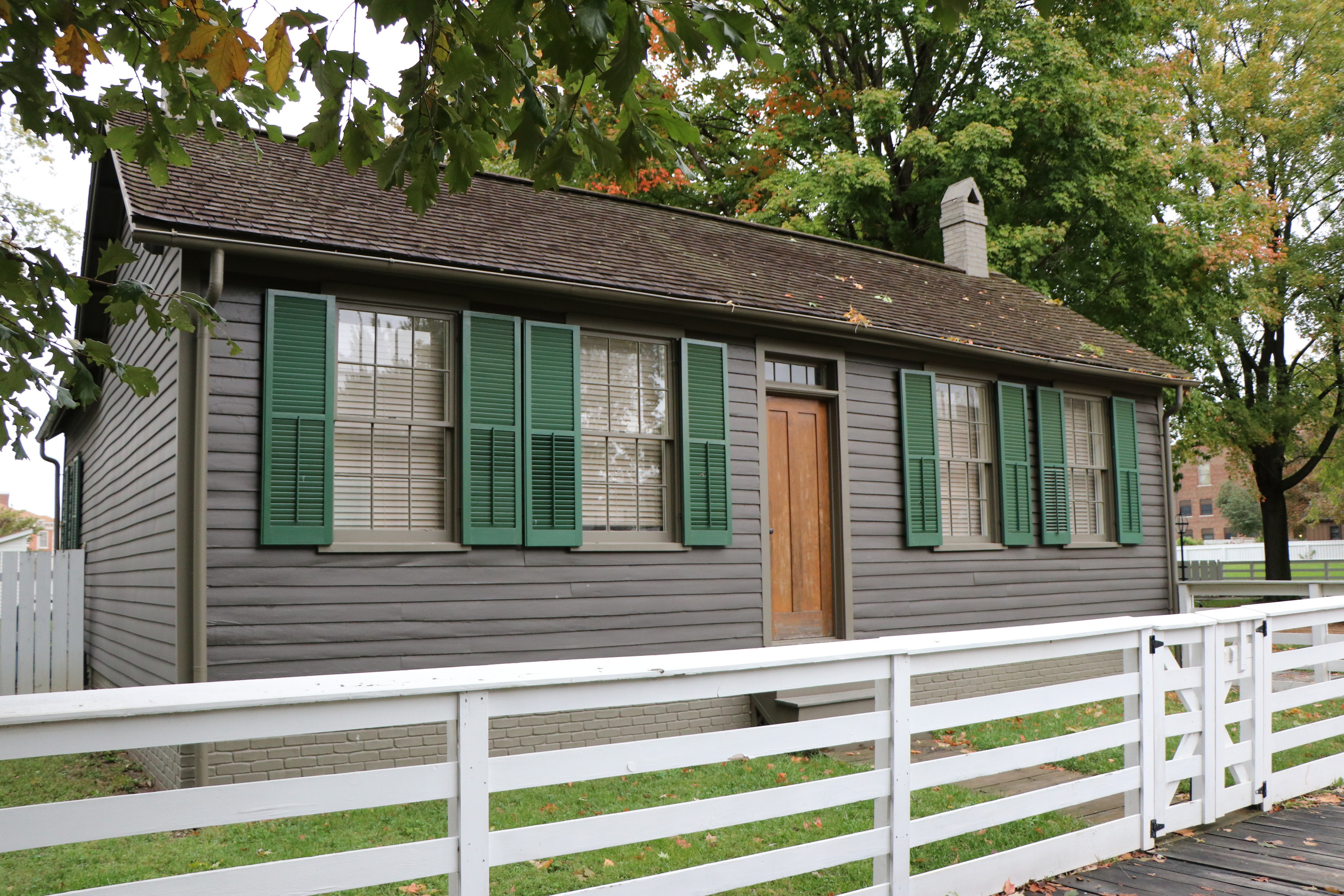 A one-story dark grey house with green shutters and a white fence in front of it.