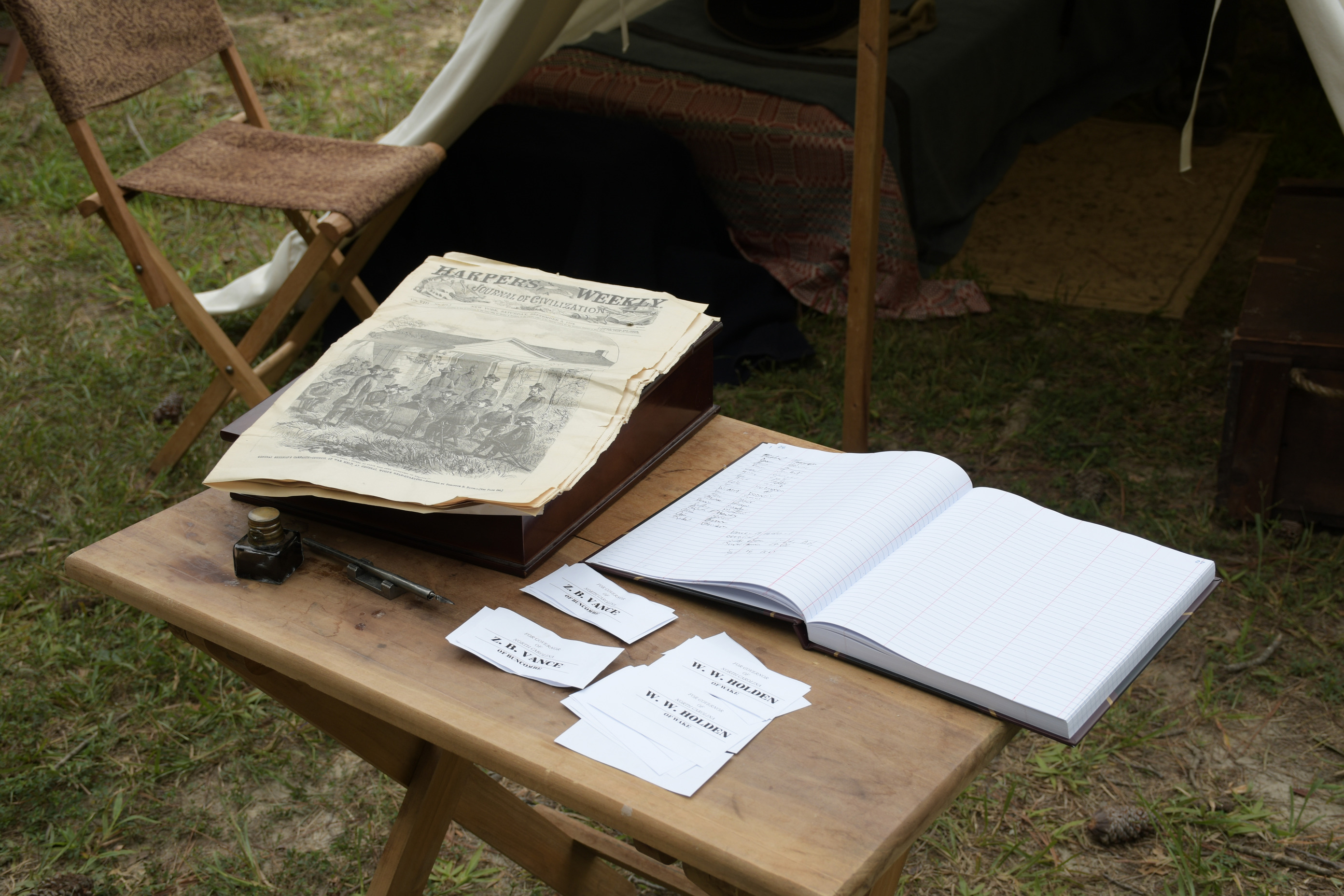 Ballots, a ledger, ink and pen, and a Harpers Weekly are placed on a wooden writing table in front of a tent. 