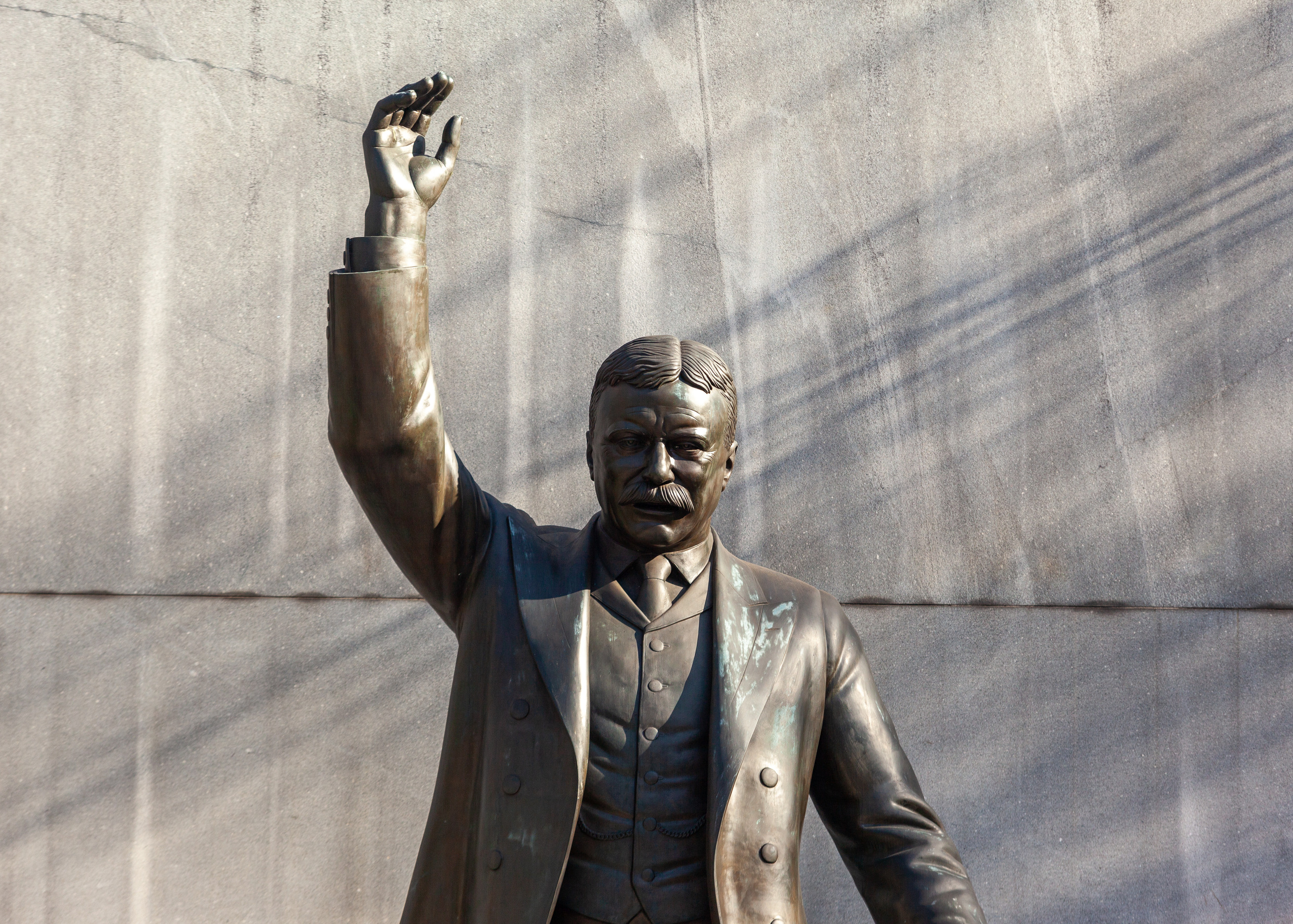 Shadows are cast behind the Theodore Roosevelt Memorial; in which Theodore Roosevelt has his right hand raised and appears to be speaking.