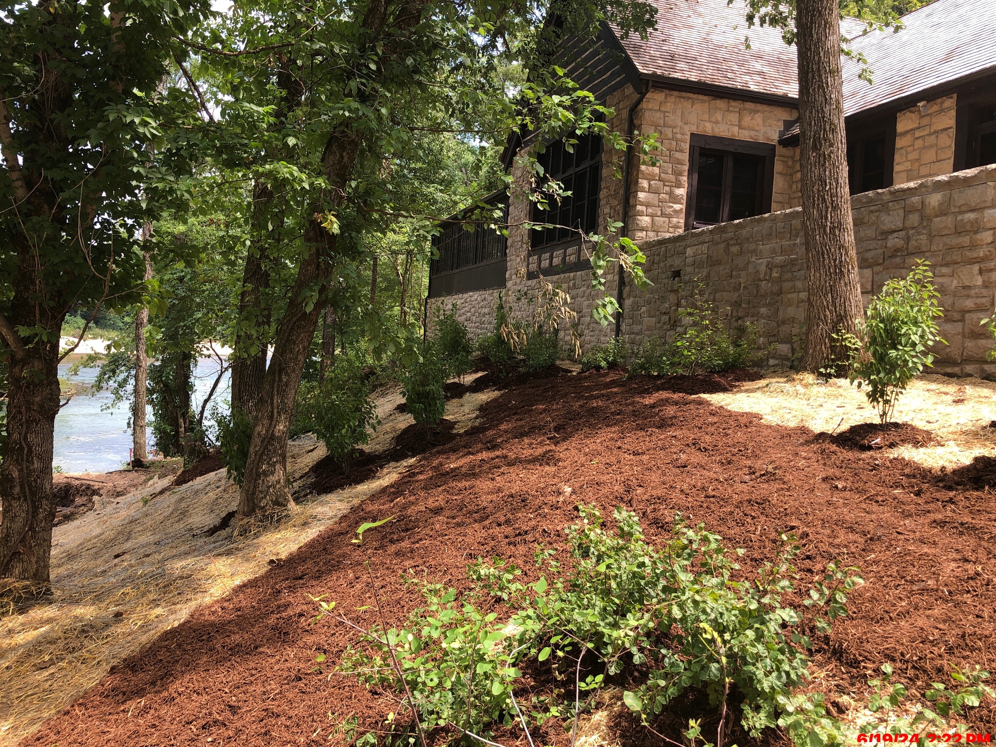 New landscaping and mulch are being installed on a shady hillside in front of a stone building with wood framing and a peaked wood shingle roof that faces to the left from the right side of the frame.