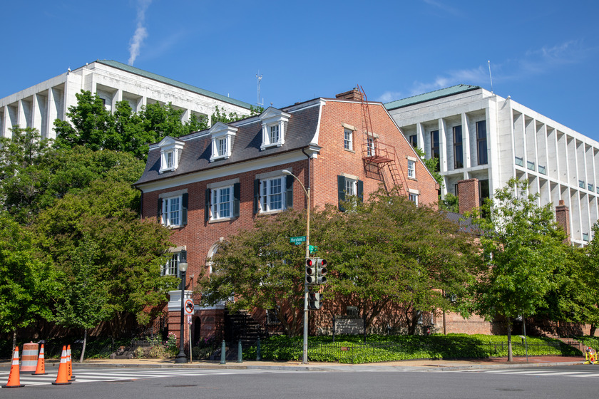 A four-story brick building on the corner of a intersection surrounded by trees and a tall white building.