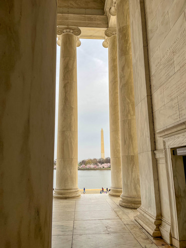 View from in between tall classical-style columns looking out across a tidal basin with a line of cherry blossom trees and a tall obelisk-shaped monument 
