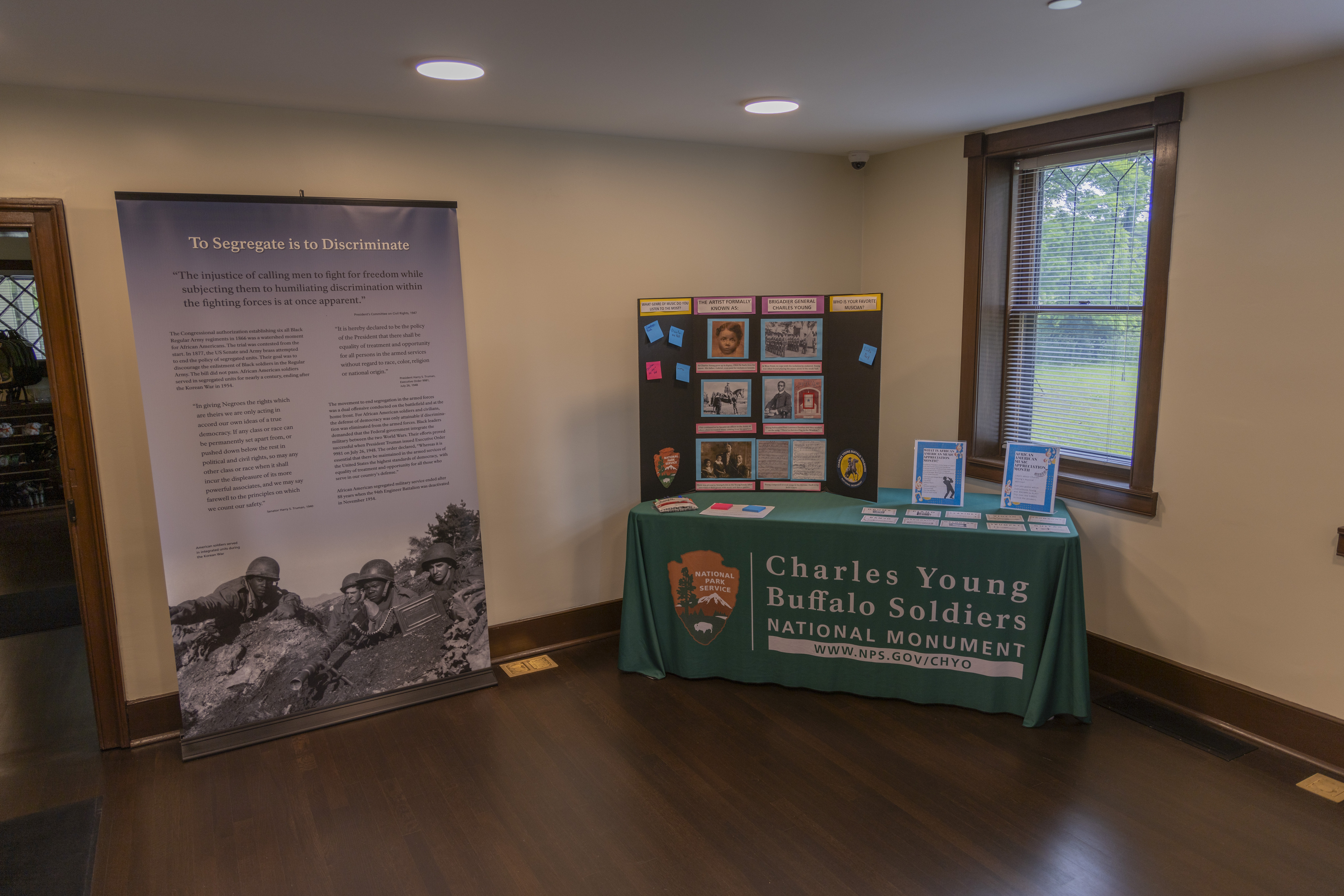 A light blue-colored pull-up banner with text and photos on them next to a table with a dark green tablecloth with printed text that reads Charles Young Buffalo Soldiers National Monument and NPS arrowhead to the left of the text, inside of a room in the Young family home.