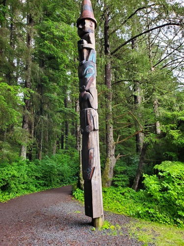 A Northwest Coast totem pole. At the bottom is  a carved fish, in the center is a wolf, and on the top is a human wearing a tall hat. 