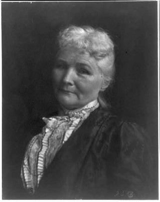 A black and white portrait of an old woman. Her face is lined, and her white hair is pulled back away from her face. Her attire is an Edwardian style, with a black jacket over her white lace covered blouse. Her expression is serious.