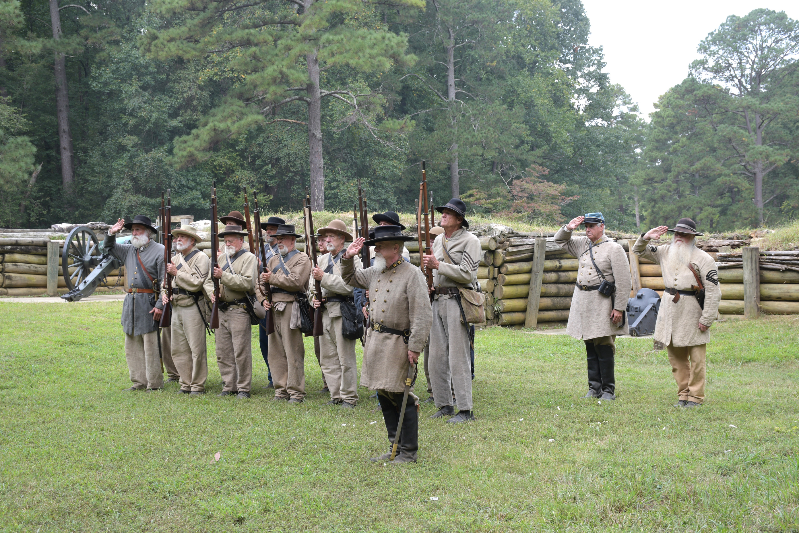 Sixteen white men wearing Confederate uniforms stand in formation offering a salute.