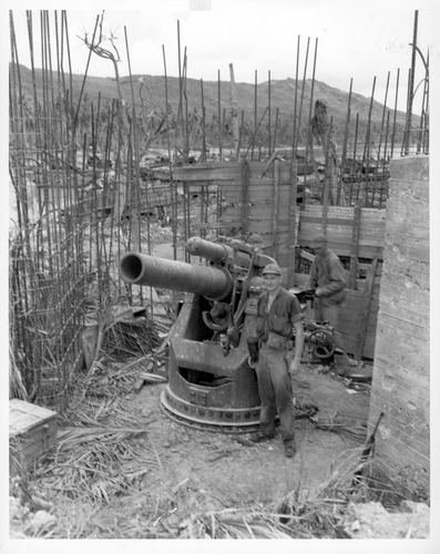 Two male soldiers in uniforms stand next to a large gun. They are surrounded an incomplete wood and rebar formwork that would be used to pour concrete into.