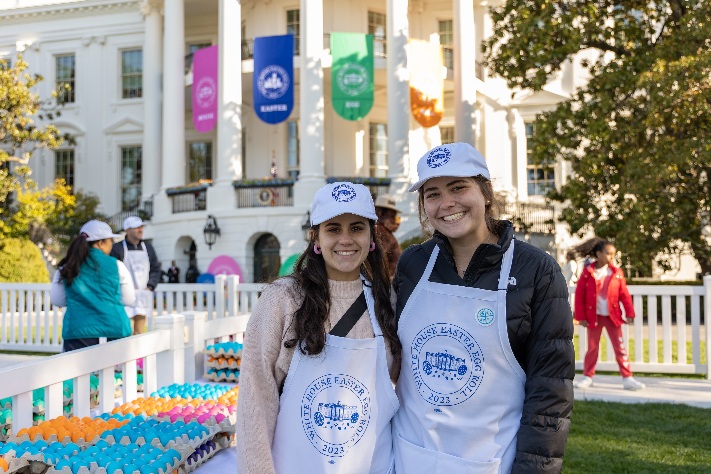 Two visitors smile with colorful Easter eggs and the white house behind them