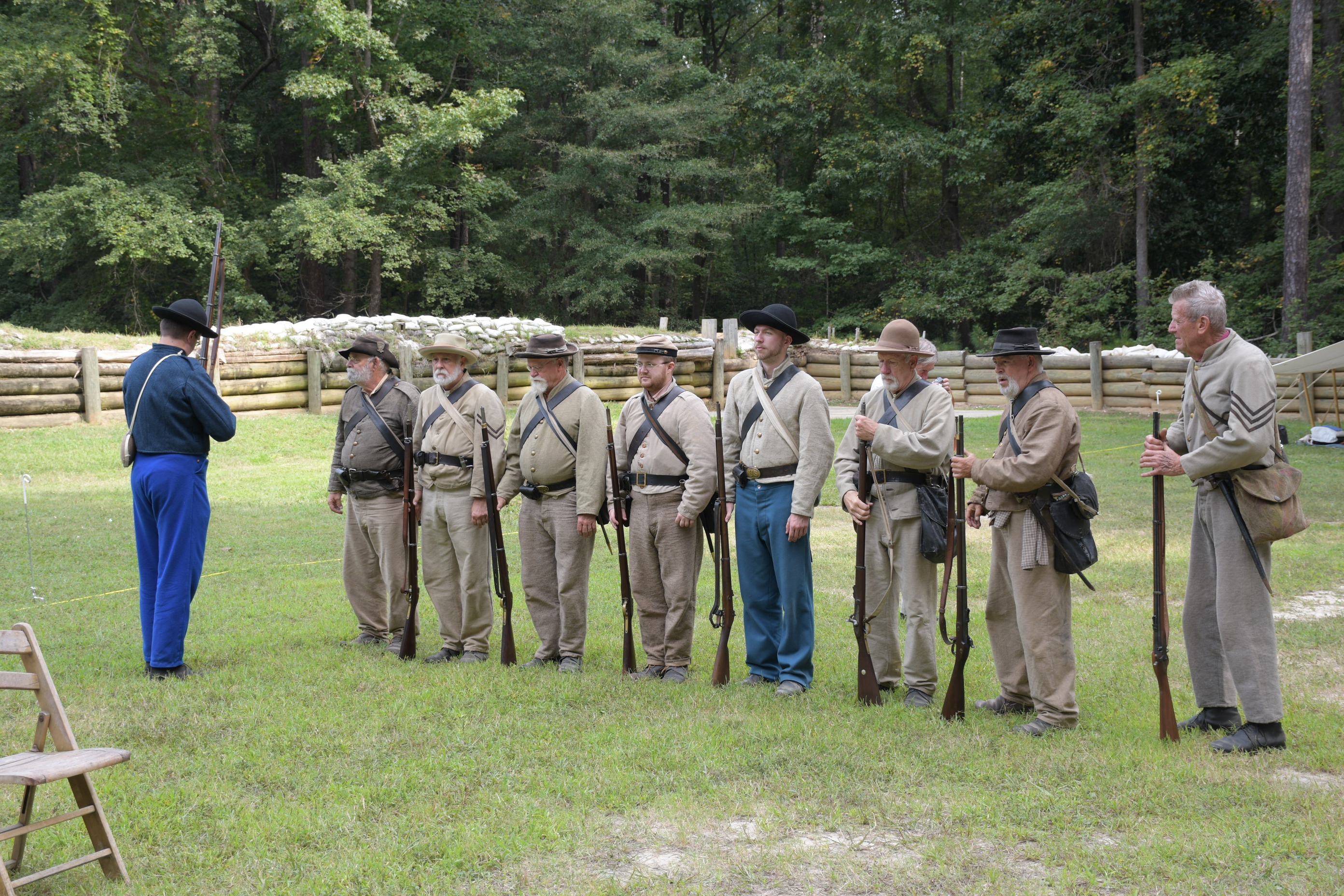 Eight white men wearing Confederate uniforms stand in a line shoulder to shoulder holding rifles. A man in wearing a Confederate uniform inspects the rifles. A wooden retaining wall supports earthworks topped with sandbags in the background.
