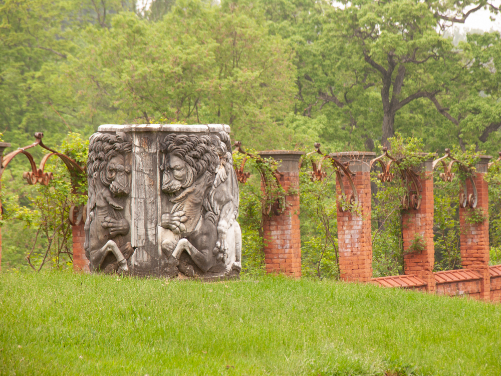 A marble wellhead with carved lions sits on a lush green lawn with brick rose trellis in the distance. 