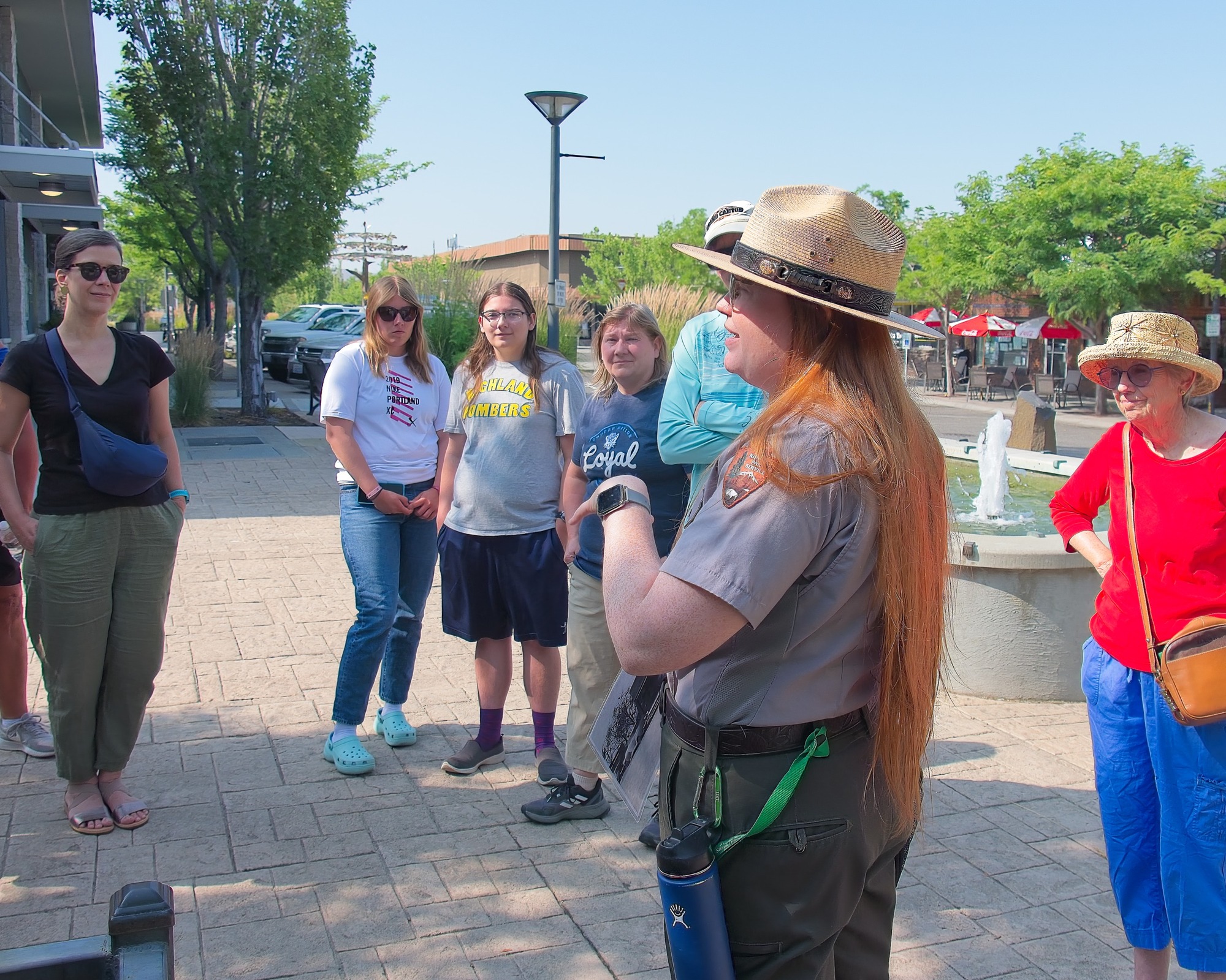 Uniformed ranger speaks to a circle of visitors standing in a downtown courtyard with trees, sunlight, and a fountain.
