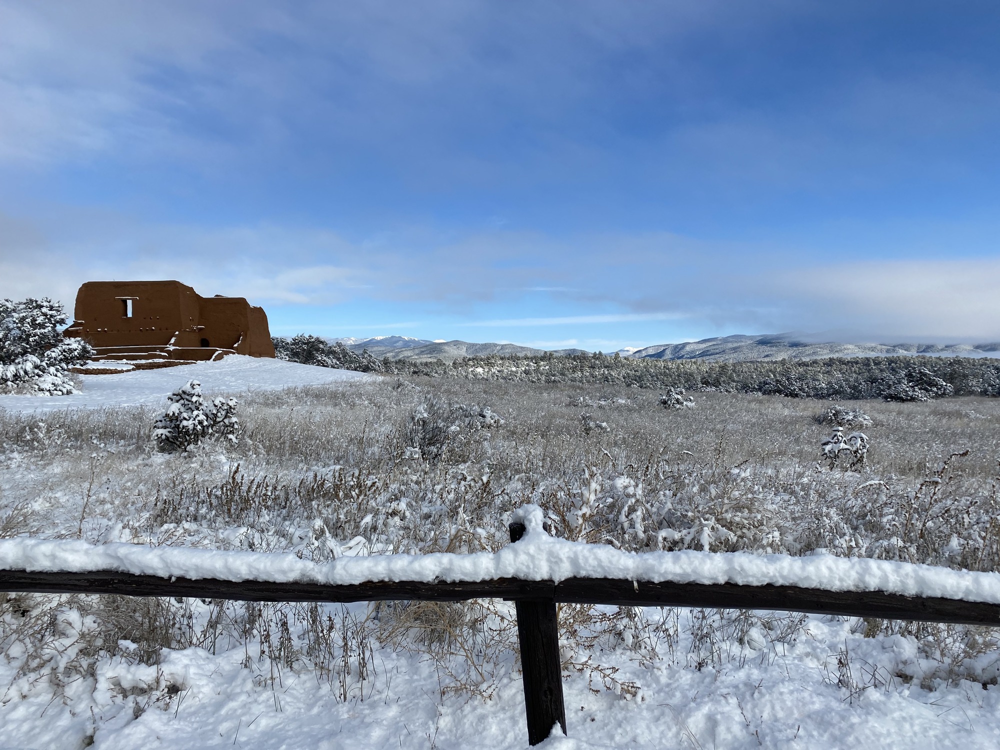 A thick blanket of white snow covers a dark brown fence that closes off a landscape view of the red adobe Spanish Mission and the snowcapped Sangre De Cristo Mountains.