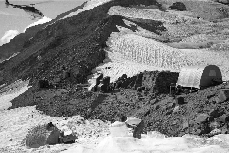 A string of small buildings along a rocky ridge, with a helicopter arriving in the upper left of the photo, and tents set up in the foreground on the snow.