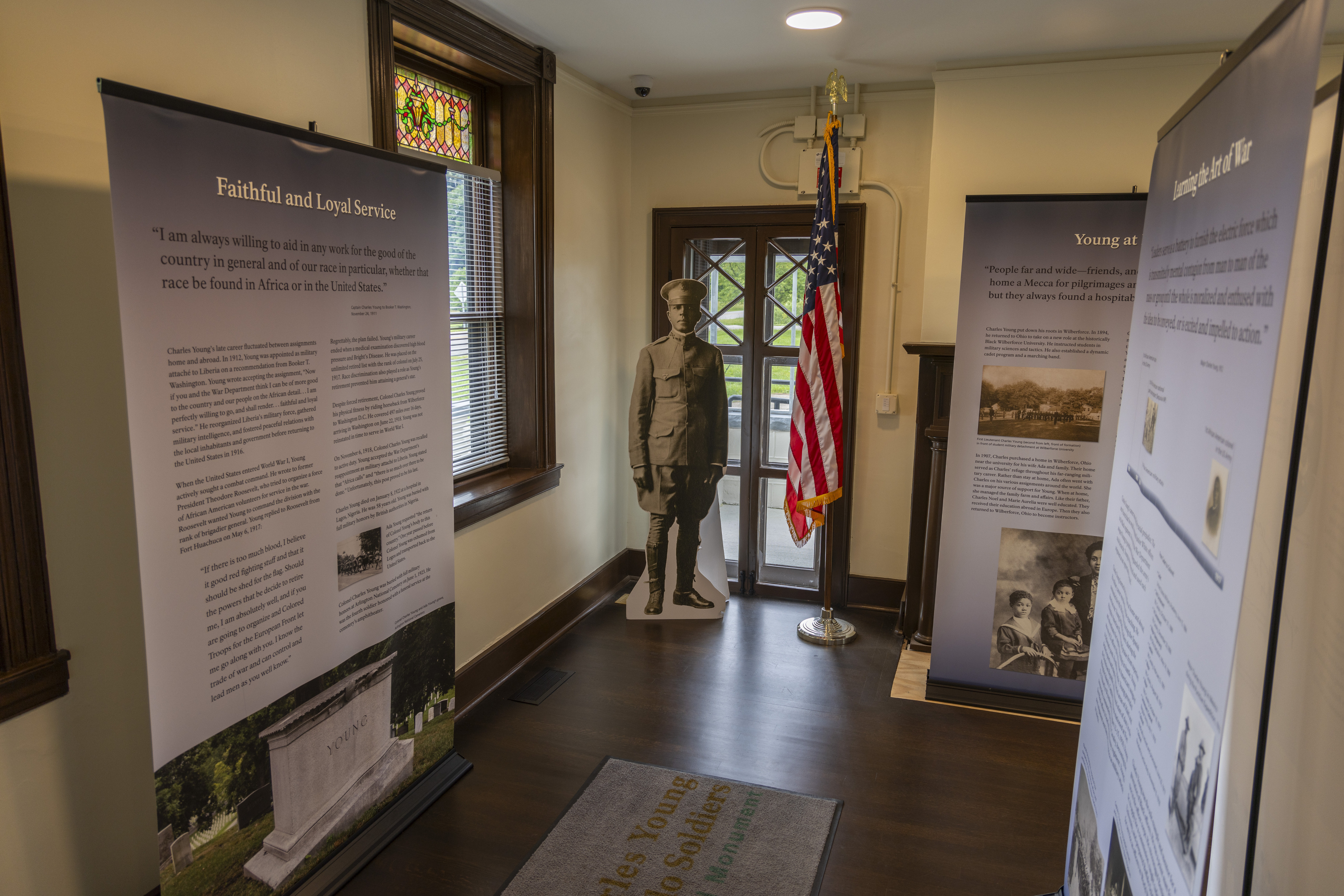 Three light blue-colored pull-up banners with text and photos on them inside of a room in the Young family home. A cardboard cutout of an African American soldier wearing a peaked cap and military uniform stands to the left of a red, white and blue American flag.