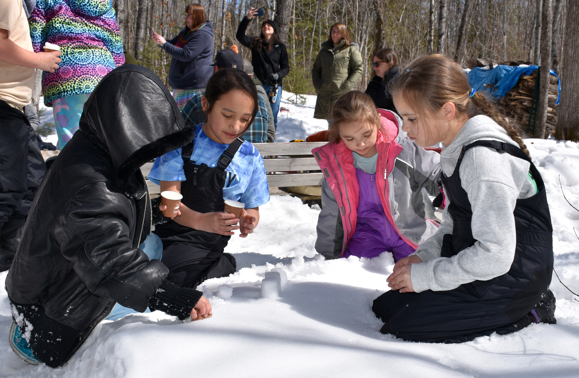 Four children use small cups to scoop and form snow to make a castle.