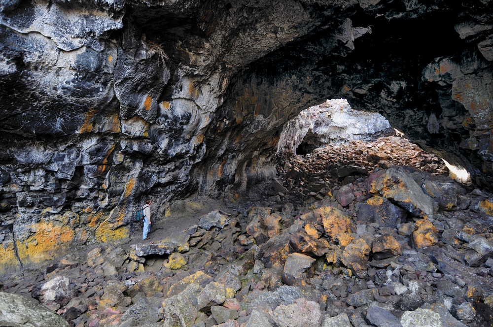 A visitor stands on a small trail along the left wall of a large cave room piled with large rocks colored with white, yellow, and orange lichen 