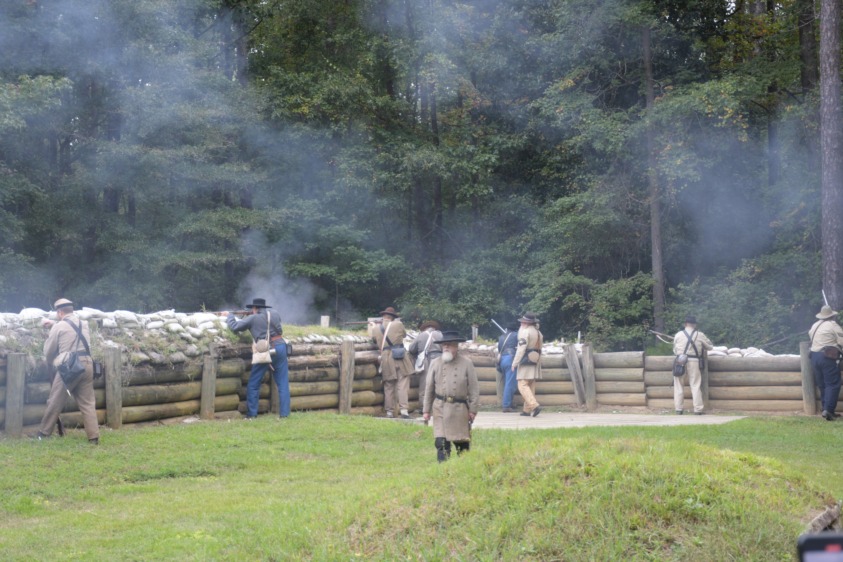 Eight white men wearing Confederate uniforms firing rifles from defensive positions behind earthworks. A white man wearing a Confederate Officer’s uniform walks among them.