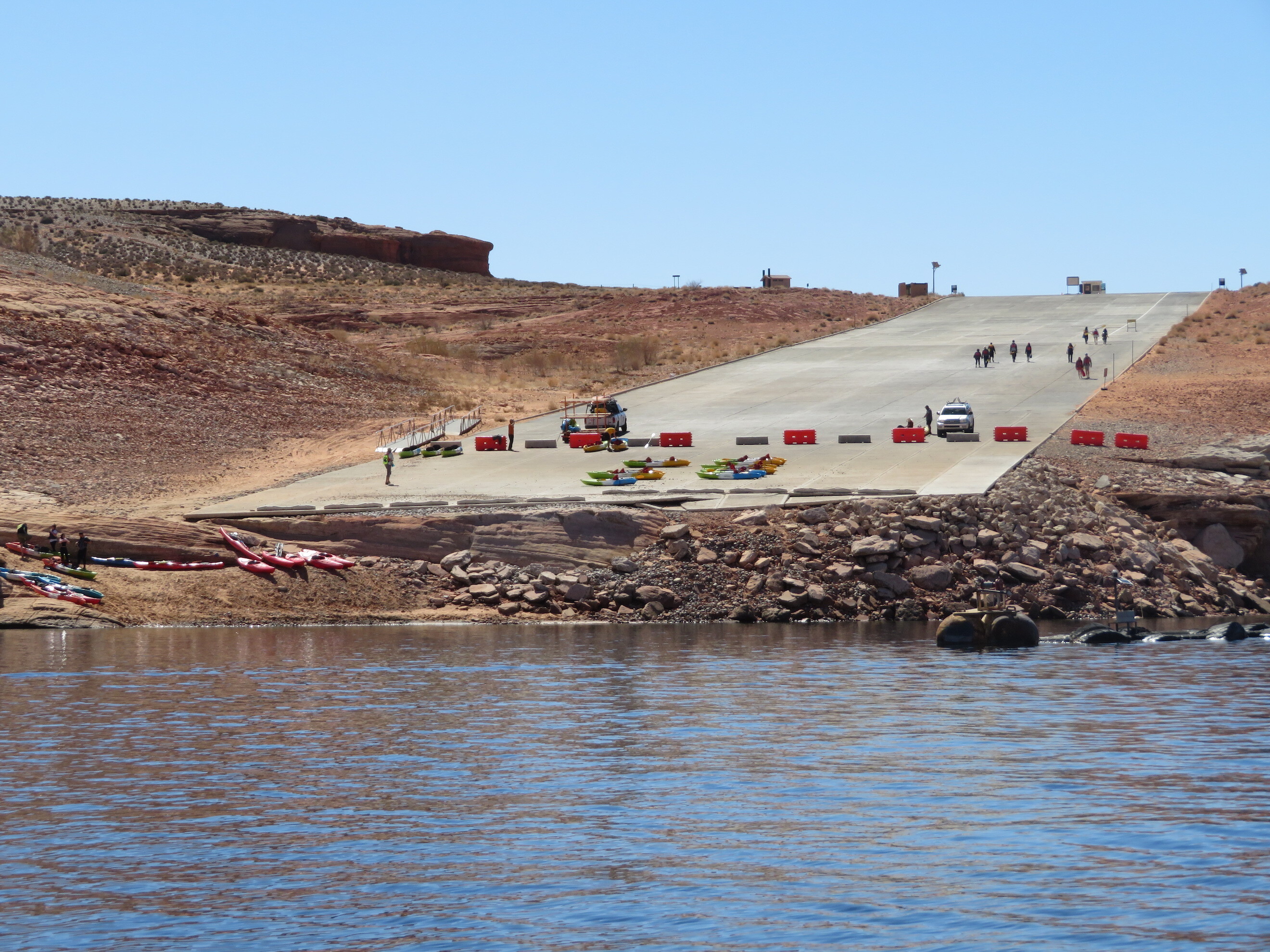 Trucks unloading kayaks on a barricaded launch ramp as visitors descend a rocky shoreline. 