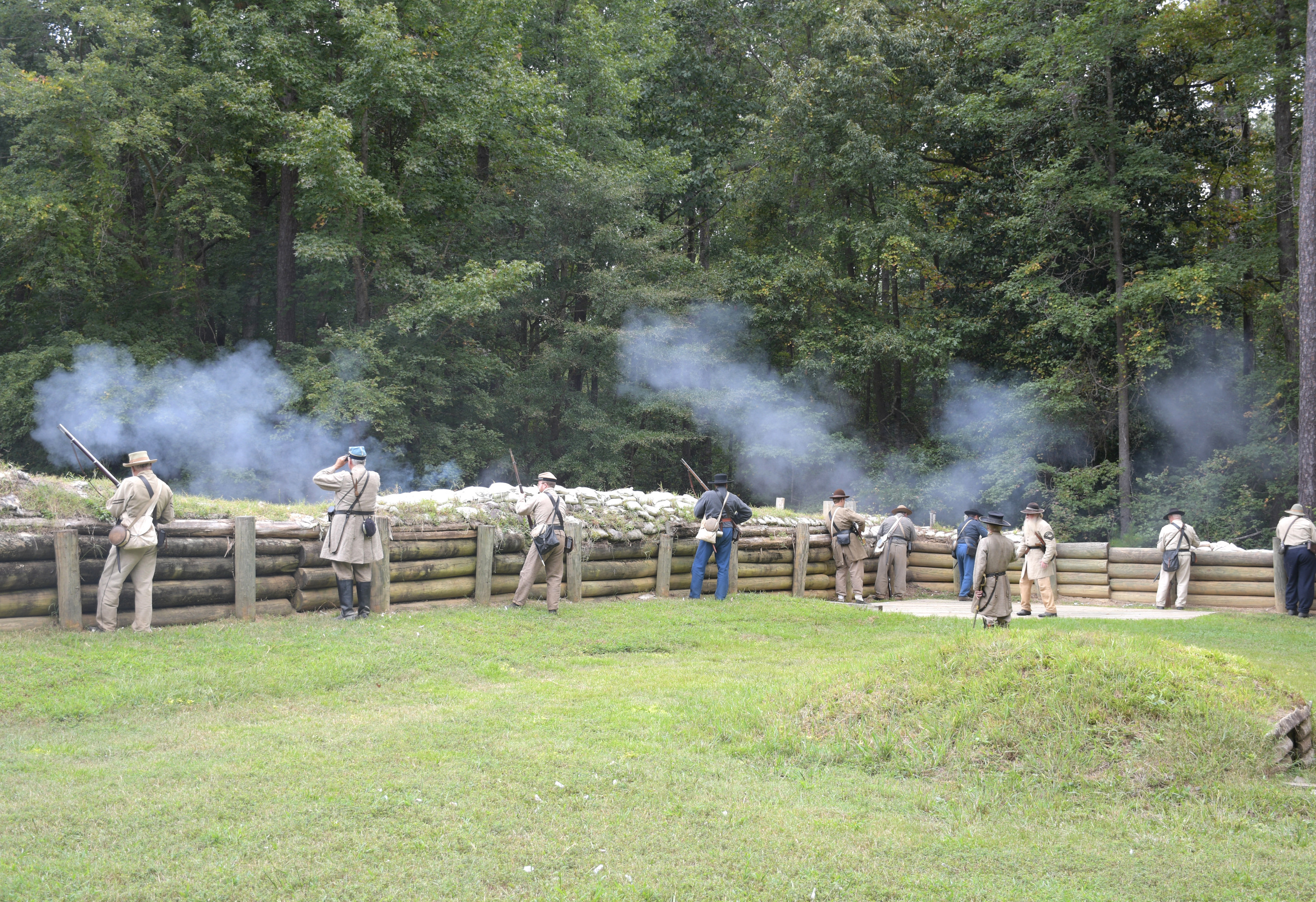 Ten white men wearing Confederate uniforms firing rifles from defensive positions behind earthworks.