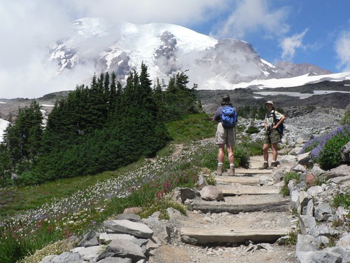 Hikers stand on a rocky trail amid subalpine meadows with Mount Rainier in the background.