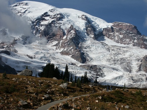 A paved trail cuts up a rocky meadow slope towards a mountain peak covered in glaciers. 