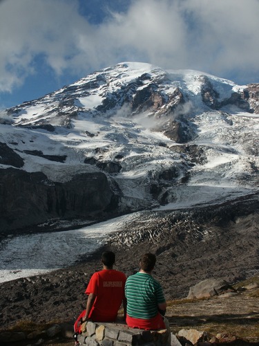 Two people sit on a low rock wall looking down on a large glacier descending a mountain peak and filling a valley. 