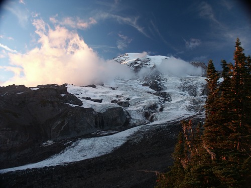 Light fades from a glacier-covered mountain peak, tinging the clouds wrapped around the summit pale pink. 